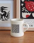 A white ceramic mug with black 'CAFFEINE' typography sits on a wooden table in front of framed wall art.