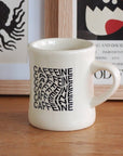 A white ceramic mug with black 'CAFFEINE' typography sits on a wooden table in front of framed wall art.