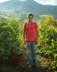 A man wearing a red polo shirt and blue jeans stands on a farm path surrounded by lush green plants, with mountains in the background under a cloudy sky.