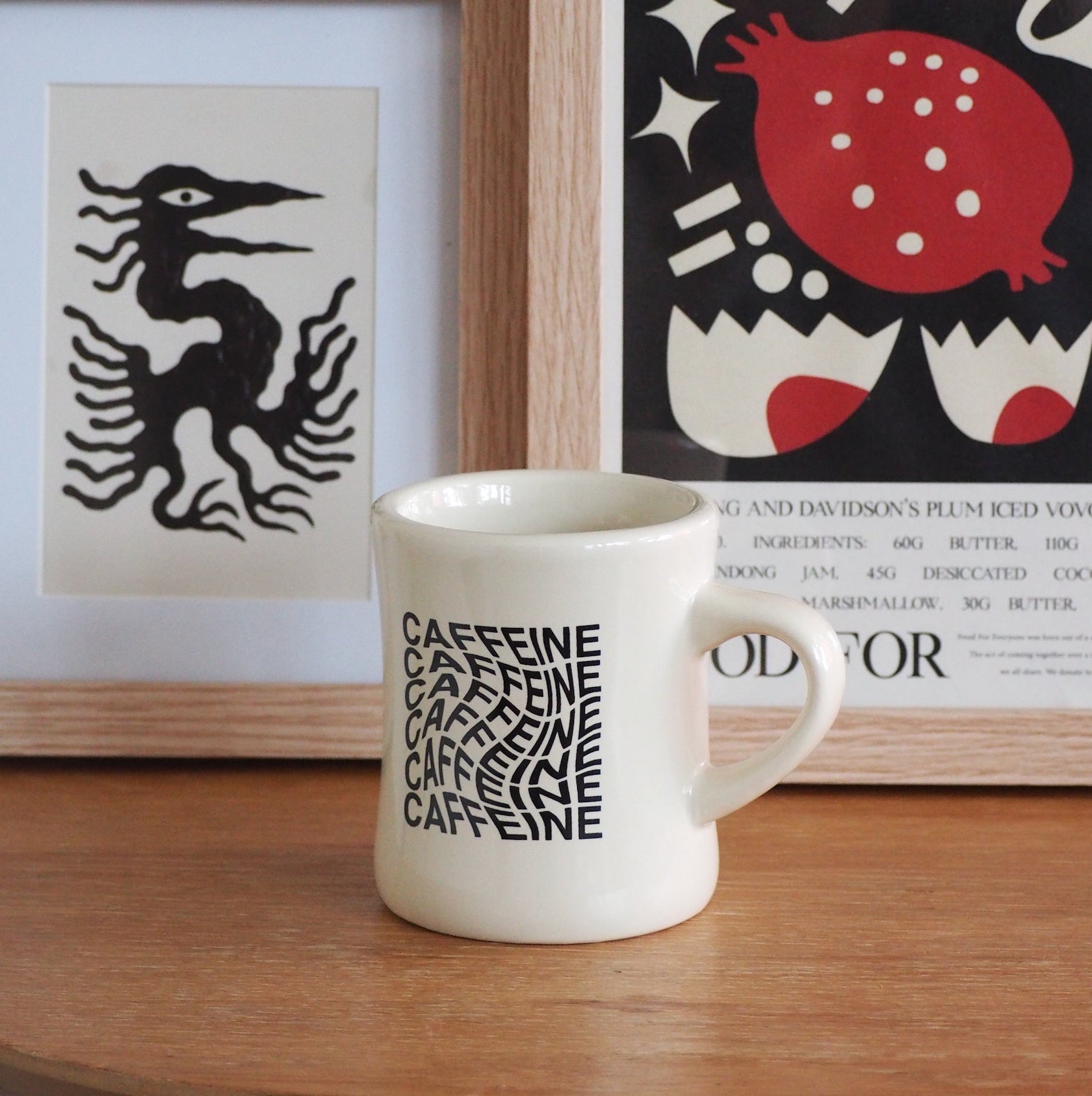 A white ceramic mug with black 'CAFFEINE' typography sits on a wooden table in front of framed wall art.