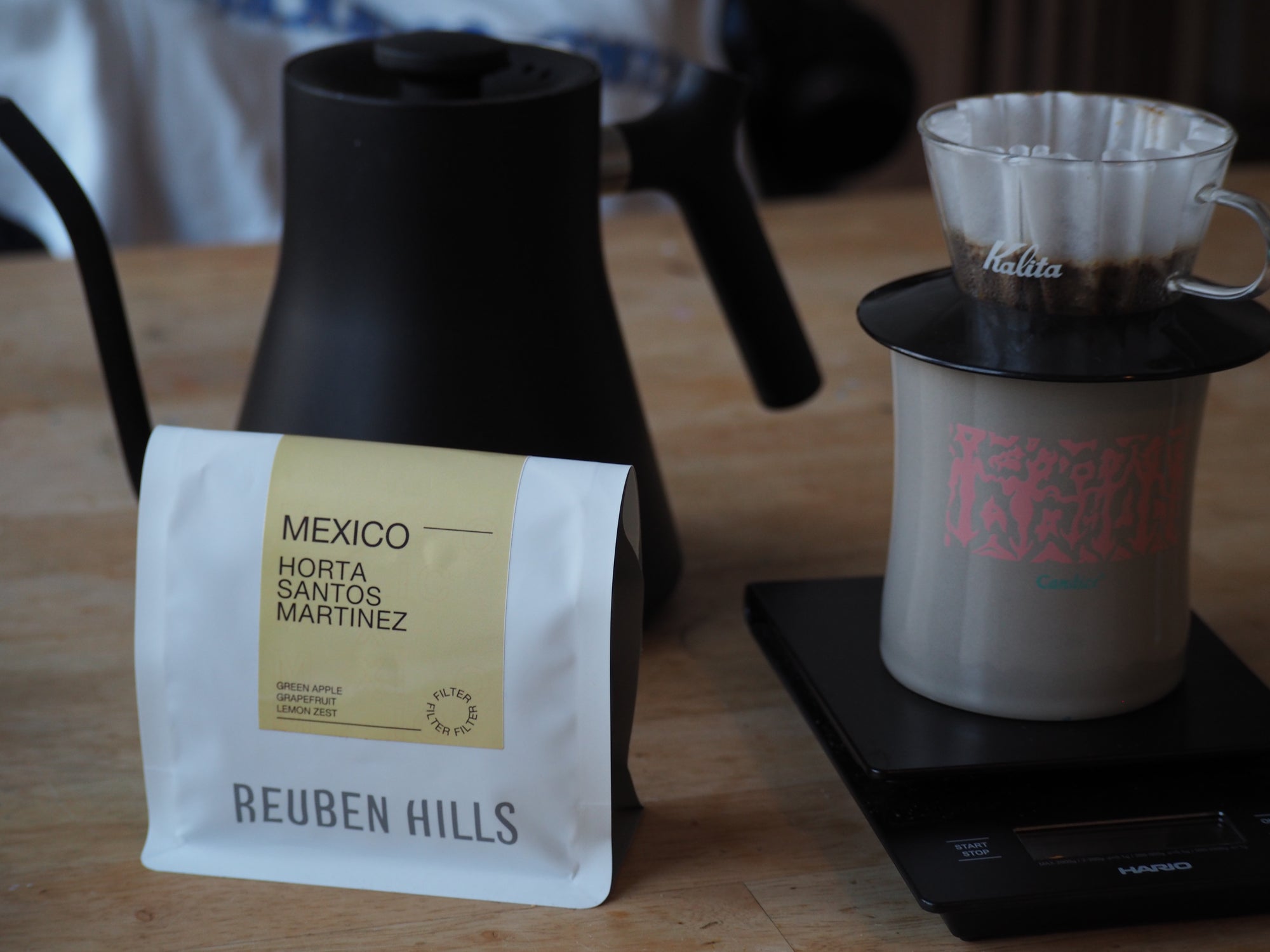 Coffee bag labeled 'Reuben Hills' next to a coffee maker and filter on a wooden surface.